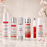 Six GYMSEGBE skincare bottles arranged on a reflective marble surface with decorative glassware and pink blossoms. The products include a white cleanser, a clear toner with silver cap, a silver serum, a red barrier-first serum, a white hydrating cream, and a white sunscreen spray with a black cap.